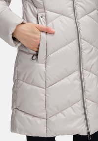 Ljusgrå vadderad jacka med chevron-quiltmönster, som har dragkedja framtill, fickor för händerna och en slät, glänsande yta.