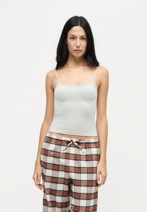 Jeune femme aux longs cheveux foncés portant un haut gris clair à fines bretelles et un pantalon de pyjama à carreaux colorés sur un fond uni.