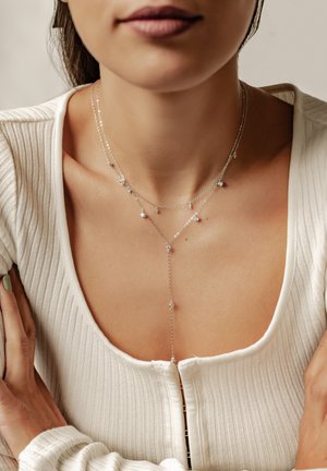 Silberne mehrschichtige Halskette mit mehreren Ketten und kleinen Edelsteindetails, getragen über einem weißen, gerippten Oberteil mit tiefem Ausschnitt.
