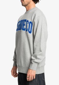 Sudadera gris con cuello redondo, que presenta un gran texto bordado azul en la parte delantera. Puños y dobladillo acanalados, material suave de mezcla de algodón.