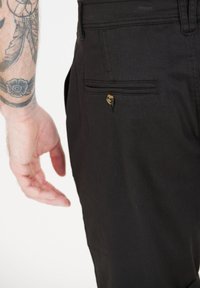 Svarta byxor med en bakficka som har en mässingsknapp. Tyget är slätt med en matt finish, synliga sömmar längs kanterna.