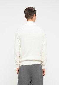 �Λευκό πλεκτό πουλόβερ με ψηλό γιακά, υφασμάτινη υφή και ραμμένα μανίκια, σε συνδυασμό με γκρι παντελόνι με φαρδιά μπατζάκια.