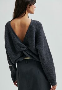 Žena s dlouhými tmavými vlasy má na sobě tmavě šedý chlupatý pletený svetr stočený na nízkém odhaleném zádech a tmavou přiléhavou sukni, stojí čelí bokem.