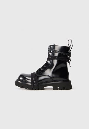 Bota de charol negro con suela gruesa, punta redondeada y detalles de cordones. Presenta cordones envueltos y una lengüeta en el talón.