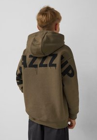 Olijfgroene hoodie met een trekkoord capuchon, met grote zwarte print op de rug en zwarte accenten op de mouwen. Zachte stof textuur.