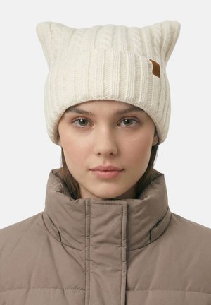 Fehér kötött sapka macskafül alakzatokkal, bordázott mintával és egy kis barna bőr címkével. Bézs színű bélelt dzsekivel viselve.