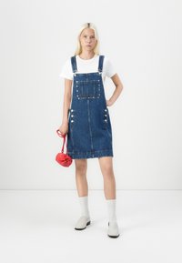Denim overall haljina s podesivim naramenicama, prednjim džepom, metalnim gumbima sa strane, uparena s bijelom majicom i crvenom torbom.