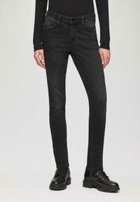 Donkerzwarte skinny jeans met een gladden denimtextuur, vijfzakkenontwerp en een klassieke knoopsluiting. Draagt met zwarte veterschoenen.