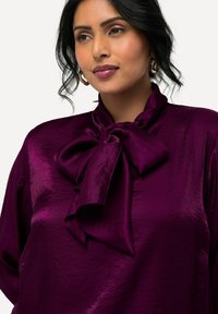 Blusa di raso viola con un grande fiocco al collo, caratterizzata da una texture lucida e una vestibilità ampia. Superficie liscia, caduta elegante e morbido lucido.