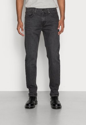 Homme portant un jean noir droit et des bottines noires, debout devant un fond uni.