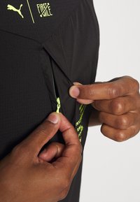 Svart idrottströja med dragkedja fram, neon gula detaljer, texturerad tyg och tryckt logotyp på sidan.