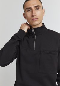 Sort fleece pullover med høj krave, halvdelslynlås foran og brystlomme. Har et struktureret stof og ribbede manchetter.