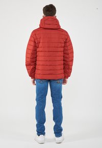 Giacca a vento rossa con cappuccio, caratterizzata da cuciture orizzontali e polsini elasticizzati. Abbinata a jeans blu e sneakers bianche.