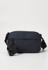 Navyblå skulderveske laget av slitesterkt stoff, med glidelåslomme foran, justerbar stropp og preget logo på forsiden.