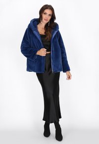 Giacca in pile blu con zip e cappuccio, morbida al tatto e dalla vestibilità ampia, abbinata a una gonna in satin nera e stivali alla caviglia.