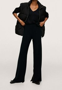 Gilet en duvet noir sur un haut sombre et un pantalon à jambes larges avec des fentes sur les côtés. Tissu lisse, design structuré et ballerines noires élégantes.