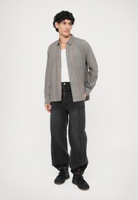 Chemise boutonnée à carreaux gris avec une poche, assortie à un jean noir à jambes larges avec des rayures latérales. Des sneakers noires complètent la tenue.