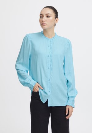 Camicia azzurro chiaro con colletto arricciato, maniche lunghe e polsini arricciati; presenta un tessuto texture e chiusura frontale con bottoni.