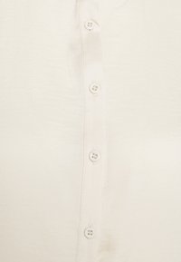Camicia abbottonata in tessuto crema chiaro, con una superficie liscia e sei bottoni a contrasto che scendono lungo il davanti.