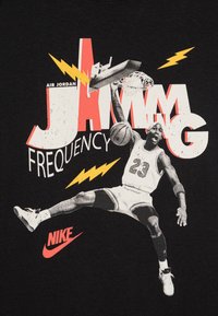 Sort t-shirt med et grafisk motiv af en basketballspiller, der hopper mod en kurv, med rød og gul tekst samt lynbolte accenter.