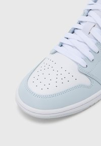 Közelkép világoskék és fehér sportcipőorról, lyukacsos mintázattal és fehér fűzővel, egyszerű háttér előtt.