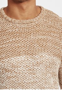 Beige och vit stickad tröja med rund halsringning, som har ett strukturerat, öppet vävmönster. Tyget ser tjockt och mysigt ut.