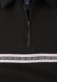 Sudadera negra con media cremallera y tejido texturizado, con una banda horizontal blanca adornada con un patrón de logotipos repetidos.