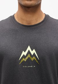 Κοντινό πλάνο ενός ατόμου που φοράει σκούρο γκρι T-shirt με διπλό λογότυπο βουνού και την επιγραφή "Columbia" στο κέντρο του στήθους.