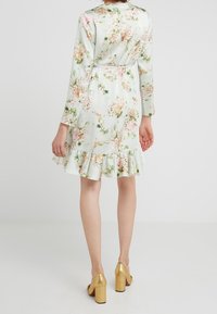 Abito floreale con maniche lunghe, tessuto di raso verde chiaro, orlo arruffato, caratterizzato da fiori rosa e bianchi. Abbinato a scarpe con tacco a blocco dorate.