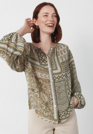 Kvinde med skulderlangt rødt hår iført mønstret knapbluse og beige bukser, smilende med en hånd bag hovedet.