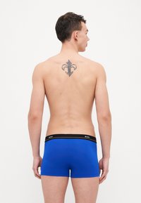 Blue boxer briefs with a black waistband labeled "BOSS." The fabric appears smooth and form-fitting. Visible tattoo on back design.