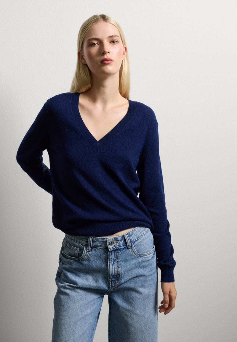 Maglione a v blu navy in tessuto morbido, abbinato a jeans blu chiaro con vestibilità classica e dettagli discreti sulle tasche.