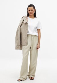 Bēša trenss, balts T-krekls un bēšas plaša kāju bikses. Gluda auduma tekstūra; sandales ar plānām siksnām. Modelis tur treniņu uz pleca.