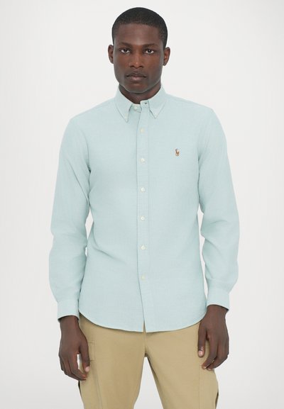 Chemise boutonnée bleu clair à manches longues, avec col et petit logo brodé sur la poitrine. Le tissu a l'air lisse avec une texture subtile.