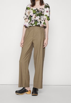Kvinde iført brune vidde bukser, en grøn blomsterprintet crop top med pufærmer og sorte træsko, stående mod en hvid baggrund.