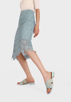 Donna che indossa una gonna di pizzo blu fino al ginocchio e sandali blu a fiori con punta aperta, passeggiando, ritagliata dalla metà del busto in giù, su sfondo bianco.