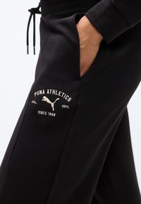 Χέρι στην τσέπη μαύρων φόρμας Puma Athletics με λευκό λογότυπο Puma και κείμενο «Puma Athletics Dept. Since 1948» στον μηρό.