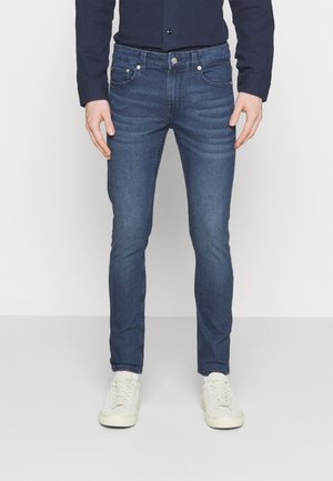 Schmal geschnittene dunkelblaue Jeans aus Denim-Stoff. Verfügt über fünf Taschen, ein dezentes Fades-Design und wird mit weißen Sneakers kombiniert.