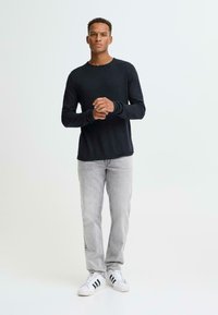 Zwart getextureerd shirt met lange mouwen gecombineerd met lichtgrijze jeans met rechte pijpen en witte sneakers met zwarte strepen.