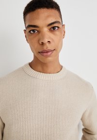 Beige gebreide trui met een gestructureerd patroon en geribbelde ronde hals, gedragen door een model met kort haar tegen een effen achtergrond.