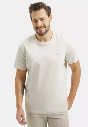 Mężczyzna z brodą uśmiechający się, ubrany w prostą, jasnobeżową koszulkę i pasujące spodnie, z tatuażem na lewym przedramieniu.