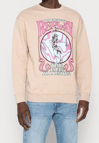 Felpa beige con una grafica colorata che presenta uno scheletro e il testo "IL TEMPO È UN'ILLUSIONE" in rosa e blu. Vestibilità standard.