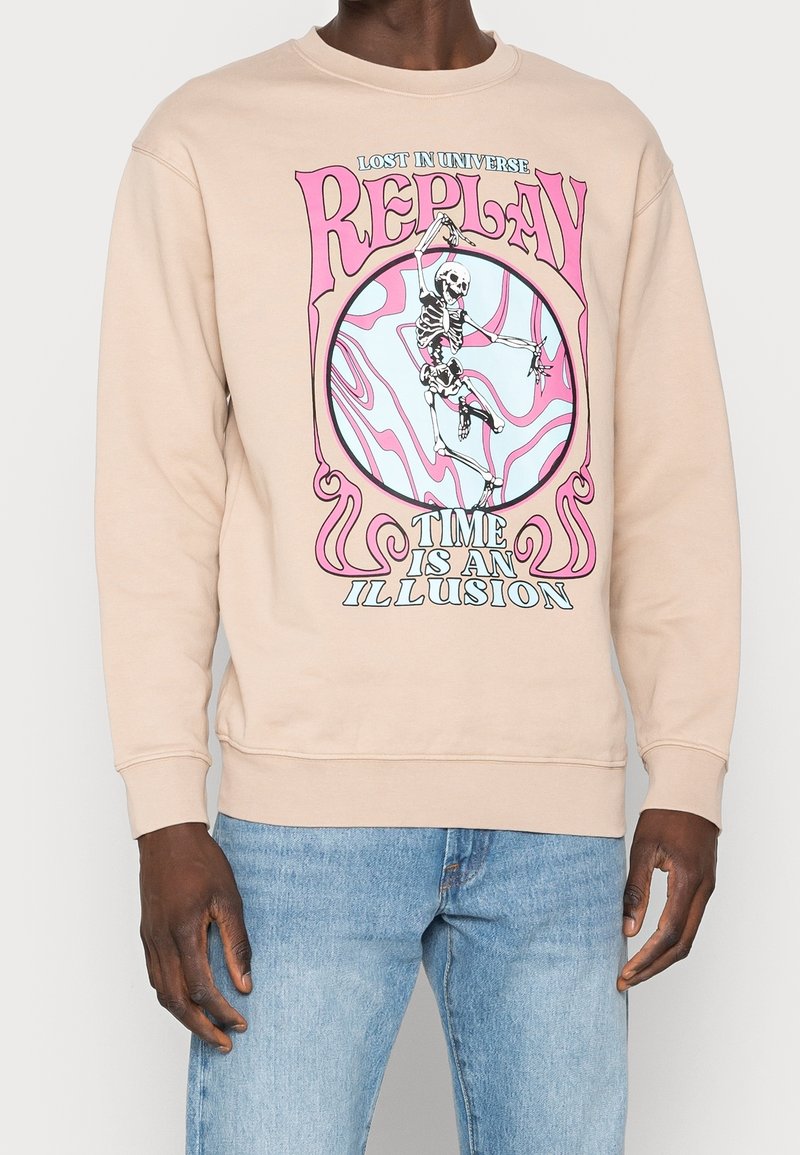 Felpa beige con una grafica colorata che presenta uno scheletro e il testo "IL TEMPO È UN'ILLUSIONE" in rosa e blu. Vestibilità standard.