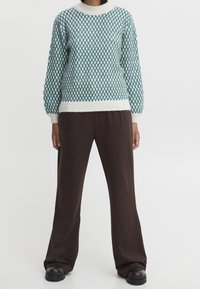 Pull en maille texturée couleur sarcelle avec accents blancs, doté d'un col rond et de manches longues. Associé à un pantalon marron à jambes larges et des chaussures noires.