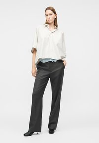 Γυναίκα που στέκεται με τα χέρια στις τσέπες, φοράει λευκό άνετο πουκάμισο με κοντά μανίκια, σκούρο παντελόνι με φαρδιά πόδια και μαύρα παπούτσια με τακούνια σε λευκό φόντο.