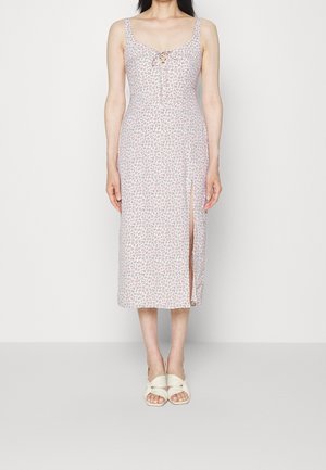 Robe midi florale en rose pastel avec une fente sur le côté, en tissu doux, avec un devant à lien ajustable et des bretelles fines. Portée avec des sandales de couleur claire.