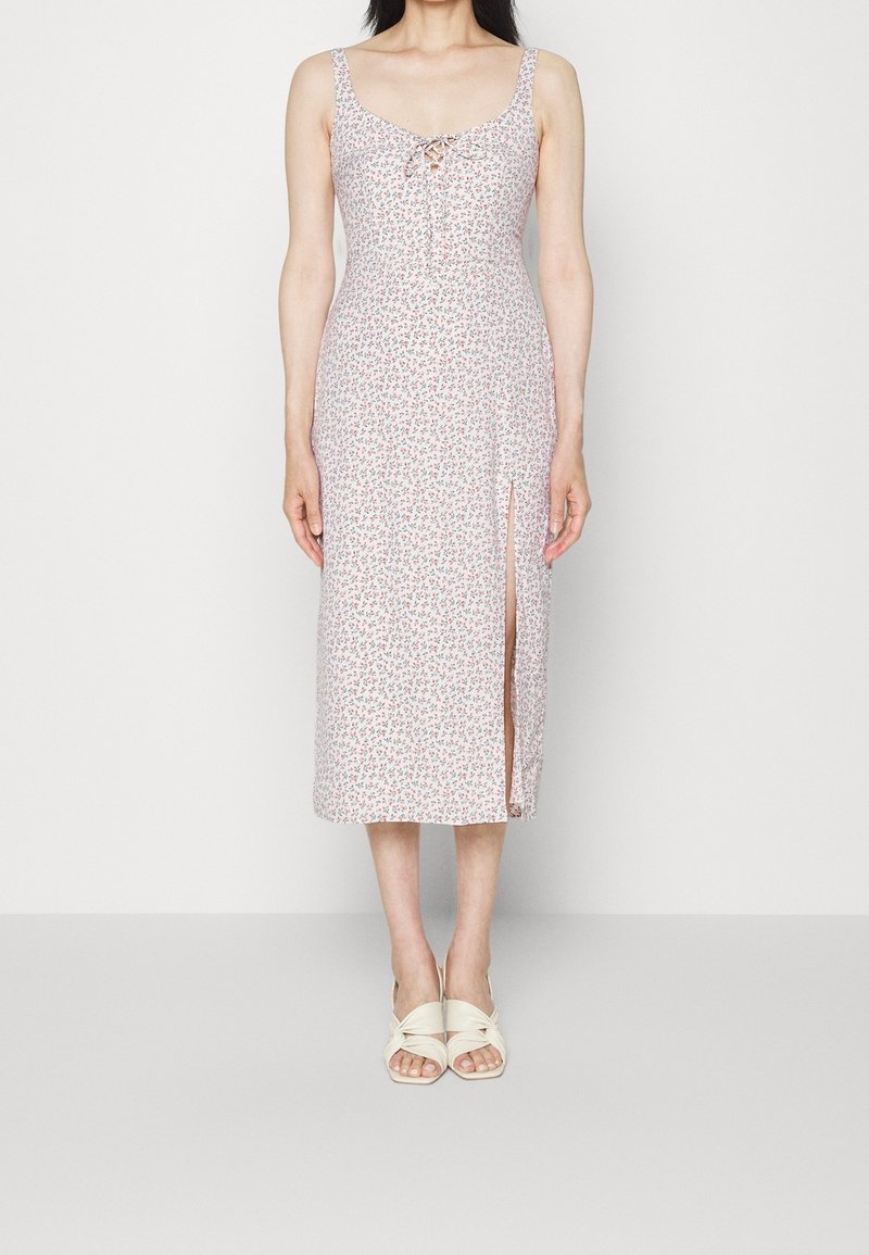 Blomstret midi-kjole i pastelfarvet pink med en sideslids, blødt stof, justerbar knytfront og tynde skulderstropper. Båret med lyse sandaler.