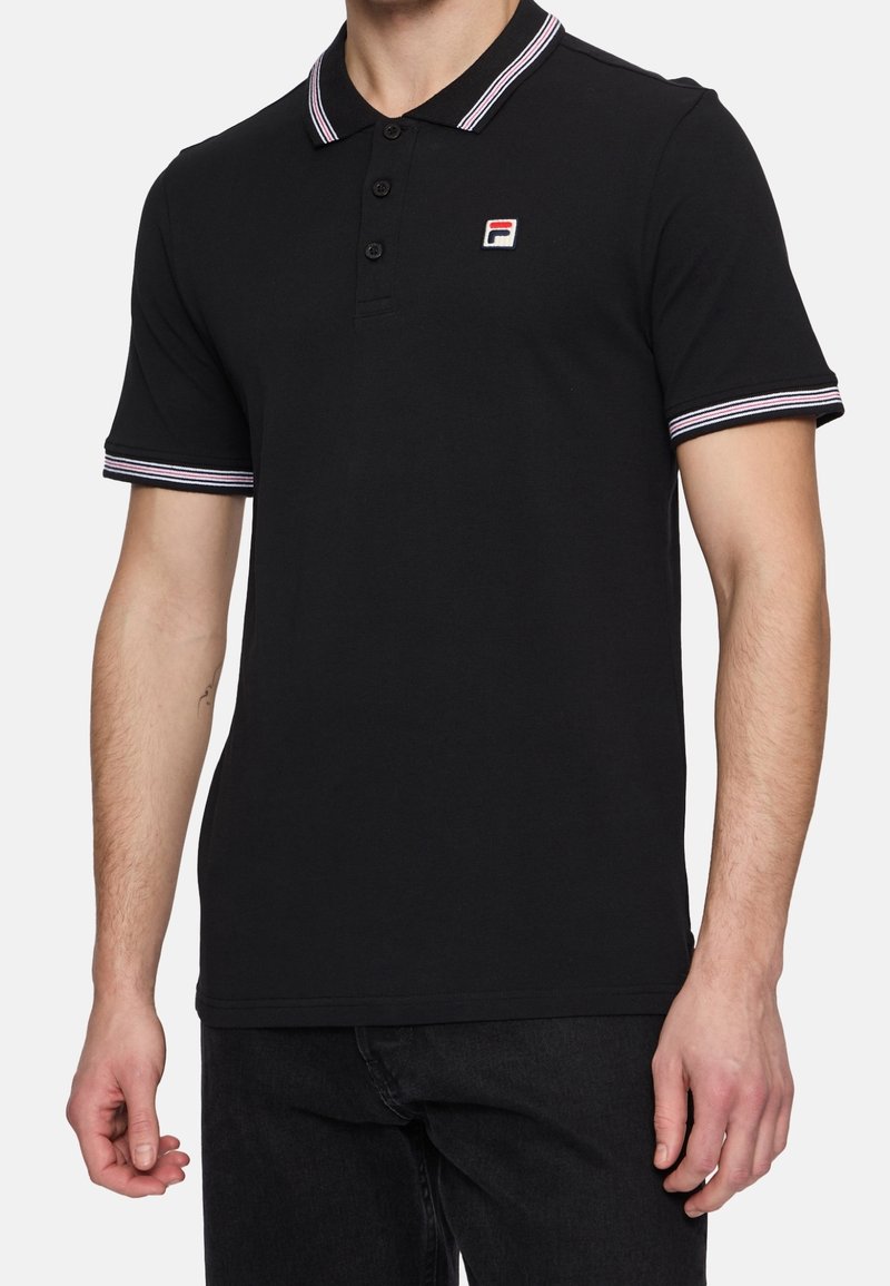 Homme portant un polo noir avec un col et des manches bordés de rayures blanches et rouges, petit logo brodé sur le côté gauche de la poitrine.