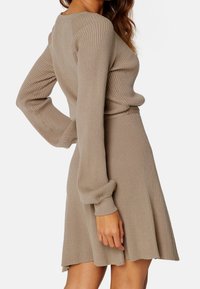 Eine Frau trägt ein beiges, geripptes Strickkleid mit langen Ärmeln und einem ausgestellt geschnittenen Rock. Sie posiert mit einer Hand auf der Hüfte vor einem weißen Hintergrund.