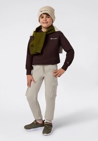 Sudadera marrón recortada con logo, pantalones cargo crema, bufanda oliva, gorro beige y zapatillas verdes con suela blanca, pose de pie.